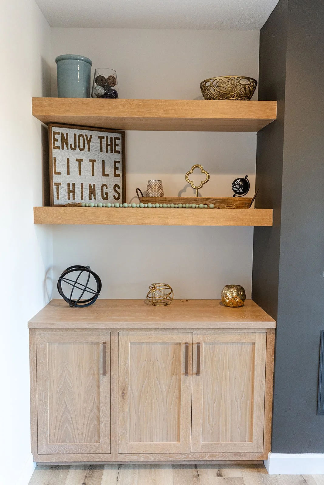 Wooden cabinet with open shelves displaying decorative items including a framed sign that says "Enjoy the Little Things," gold and ceramic bowls, a beaded necklace, metallic decorative spheres, and a small black sign.