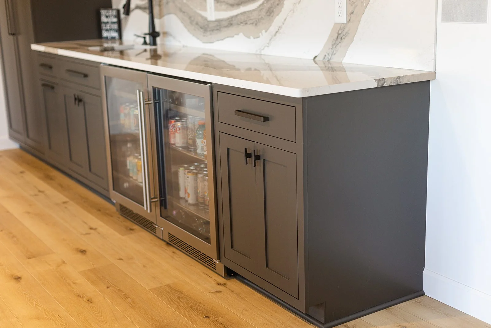 Modern kitchen bar cabinet with black and white marble top, glass door mini fridge stocked with drinks, and dark gray cabinetry, on a wooden floor.