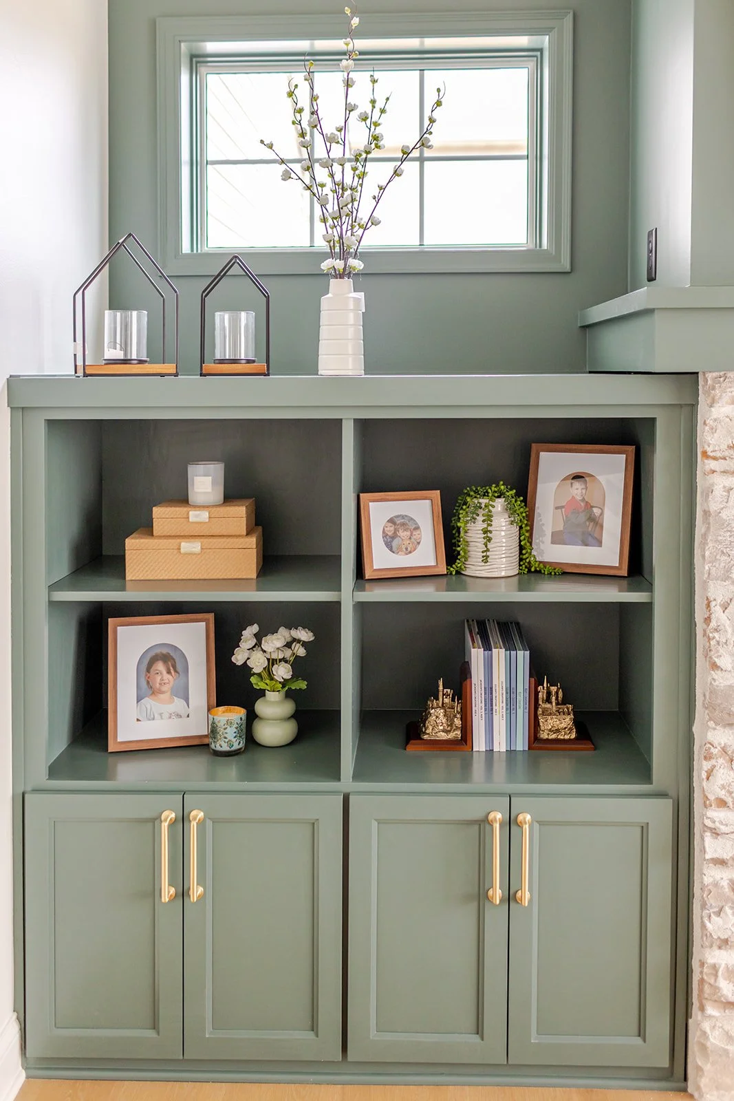 A mint green bookshelf with four shelves, decorated with framed photos, a vase with white flowers, and various decorative objects, situated against a light green wall with a window above.