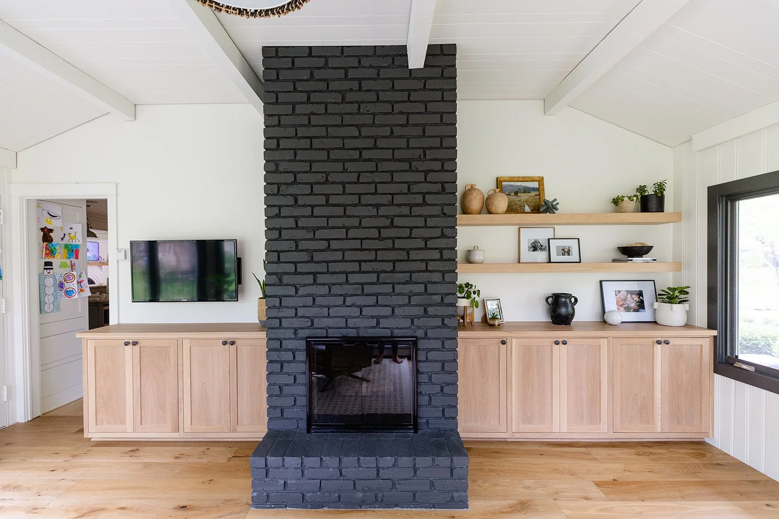 Living room with a black brick fireplace dividing two wooden media cabinets with shelves, a wall-mounted TV, and a window on the right.