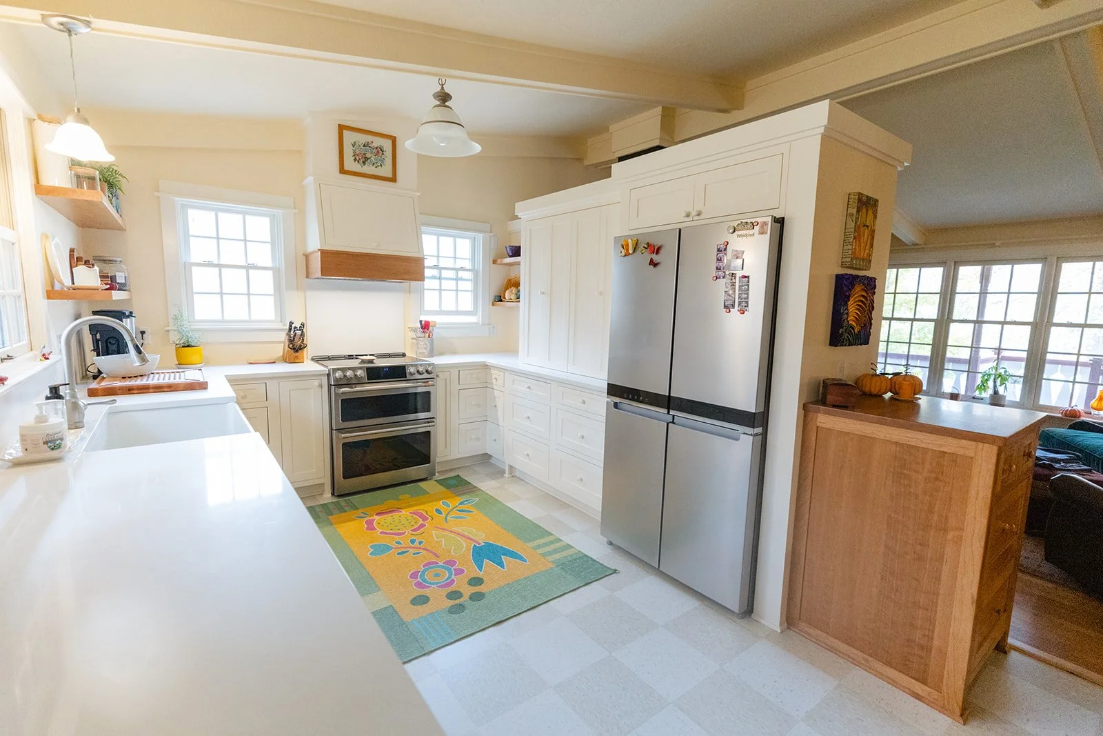 A bright farmhouse-style kitchen with white cabinetry and a colorful rug, showing a stainless steel fridge, a stove, a white countertop with a sink, and windows with natural light.