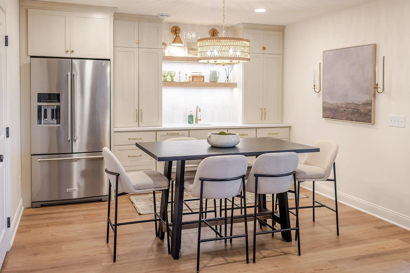 Modern kitchen with white cabinetry, stainless steel refrigerator, black dining table, six beige upholstered chairs, wooden floor, and decorative artwork on the wall.