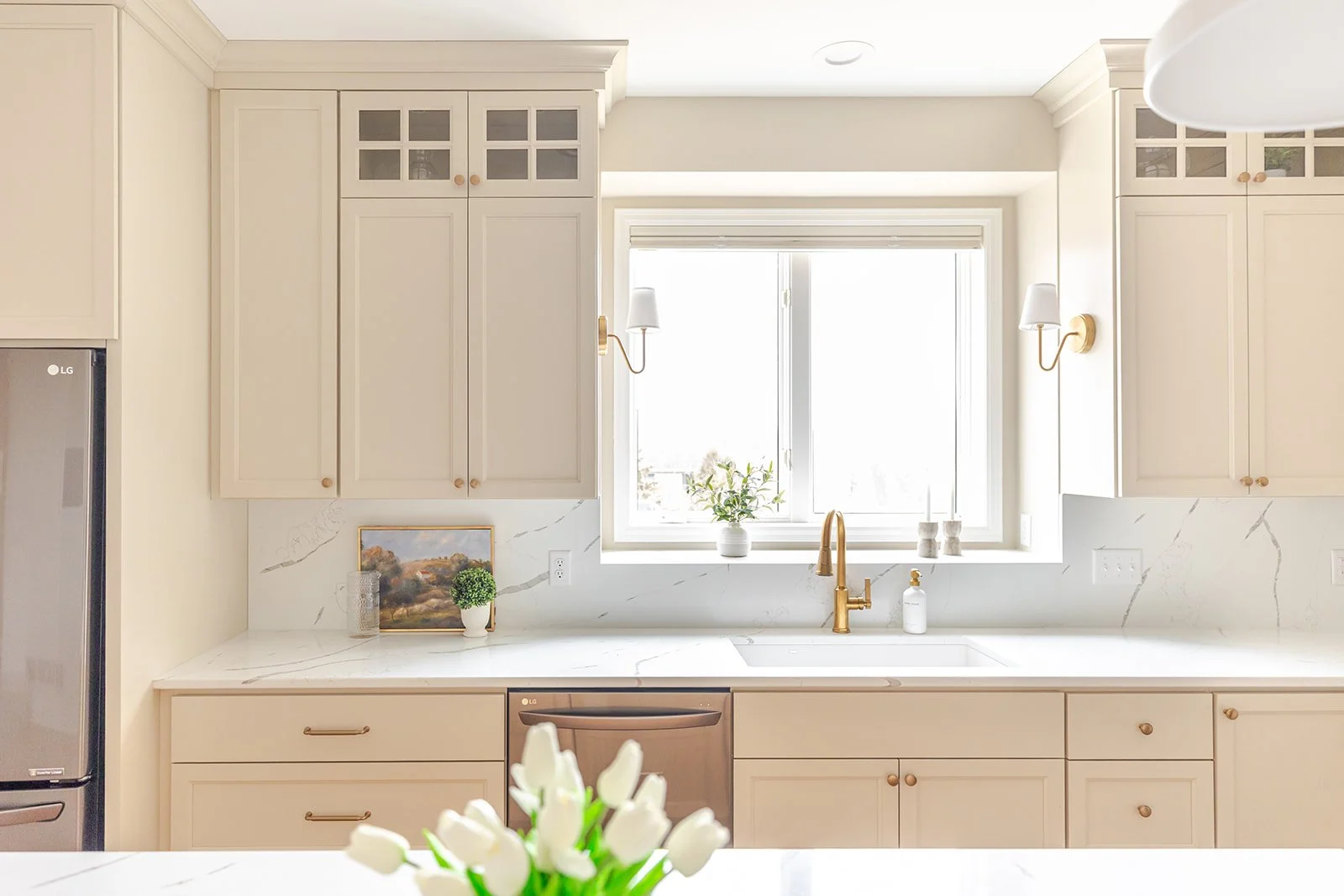Bright, modern kitchen with cream-colored cabinetry, a large window with white blinds, and gold fixtures, including a faucet. There are small decorative items on the windowsill and wall sconces flanking the window. A vase with a plant sits on the cou