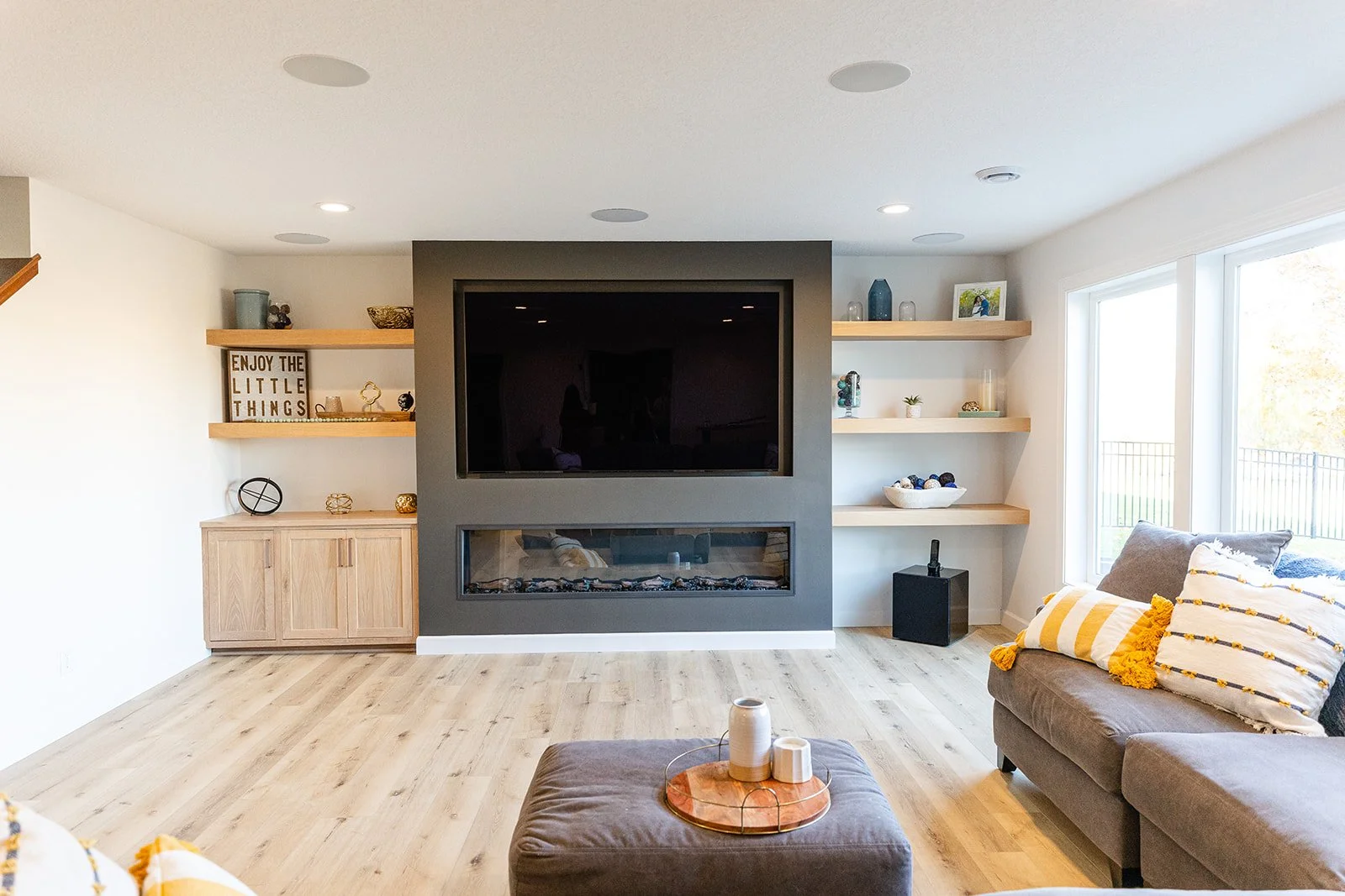 Modern living room with a gray fireplace wall, built-in shelves, a wall-mounted TV, a brown ottoman with decorative vases, and a sofa with striped pillows by large windows.