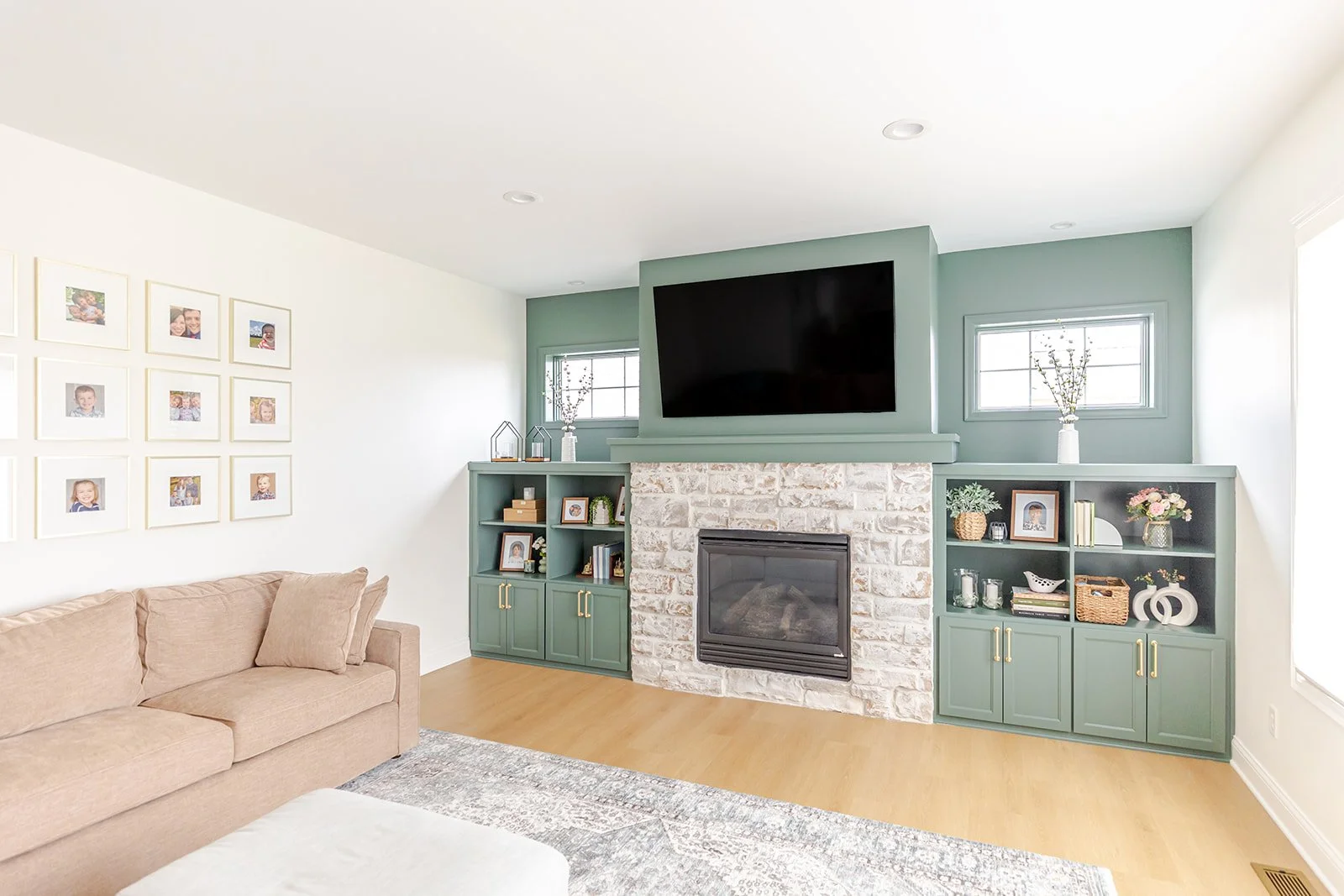 Living room with a beige sofa, picture gallery on the wall, and a fireplace with a mounted TV above. Green built-in shelves with decorations and books flanking the fireplace. Two small windows on either side of the TV, and a patterned rug on the wood
