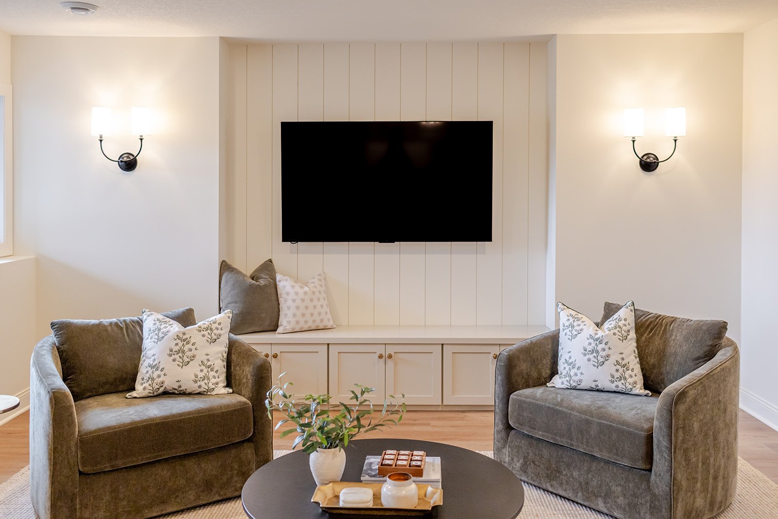 Living room with two gray armchairs, a round black coffee table with a yellow tray, pottery, and plants, a built-in white cabinet, two wall-mounted light fixtures, and a large flat-screen TV.