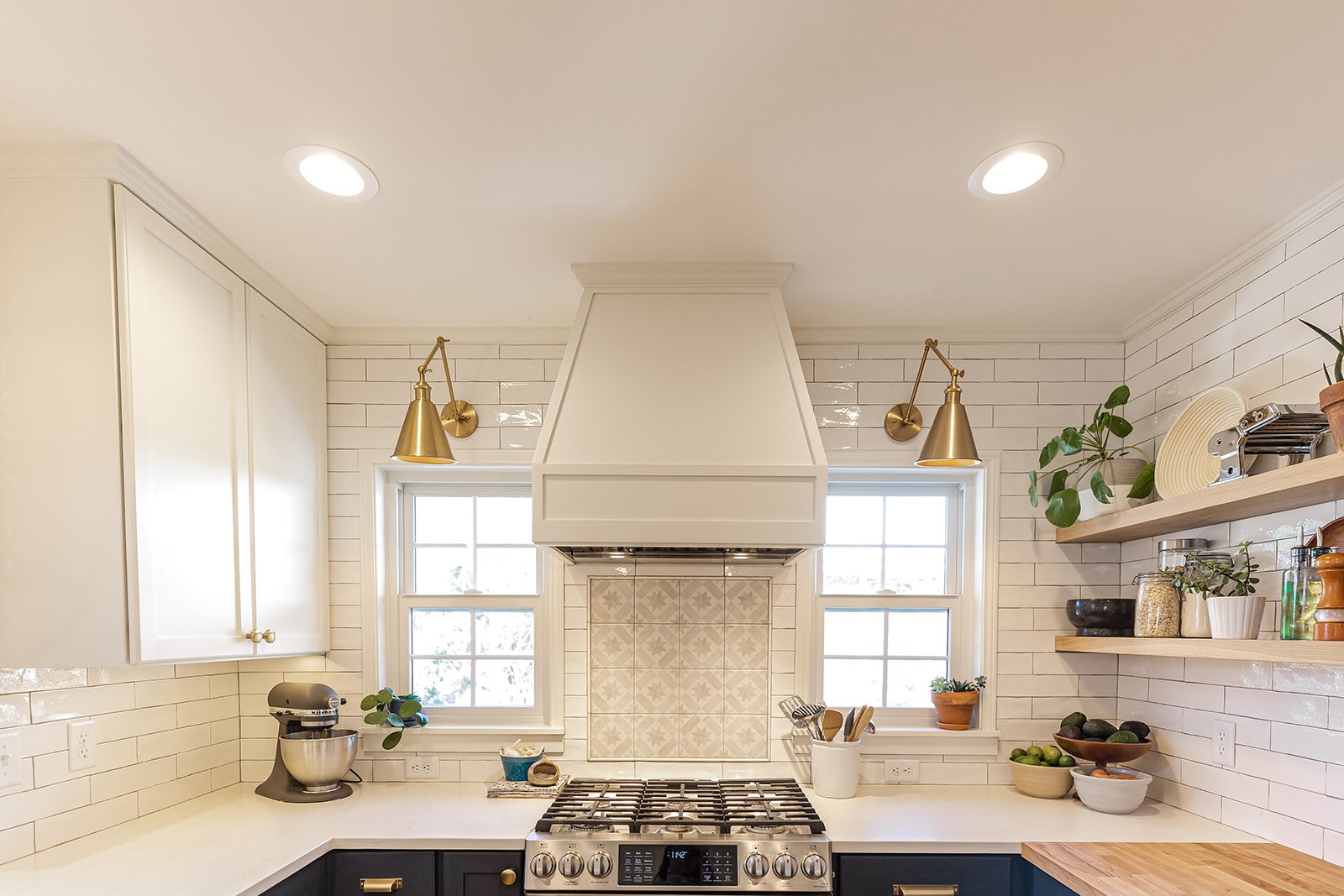 Modern kitchen with white subway tile backsplash, white cabinets, a gas stove with a white range hood, pendant lights, and open wooden shelves with plants and kitchen items.