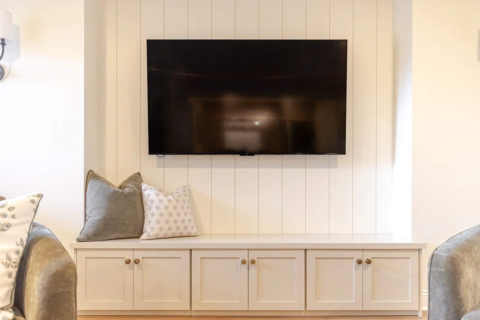 Living room with a wall-mounted flat-screen TV above a white storage cabinet. Two decorative pillows, one gray and one white with a pattern, rest on the cabinet's left side.