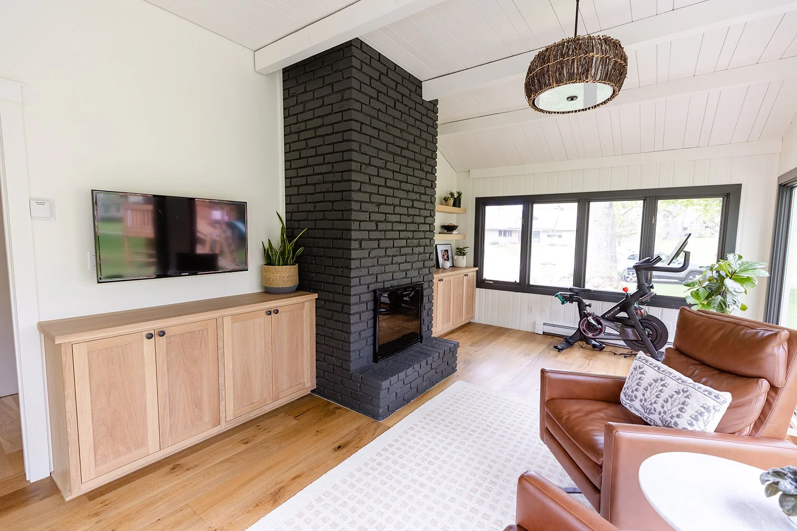 Living room with a black brick chimney, a wall-mounted television, a wooden cabinet, a potted plant, a leather armchair with a patterned pillow, a treadmill near large windows, and a ceiling light fixture.