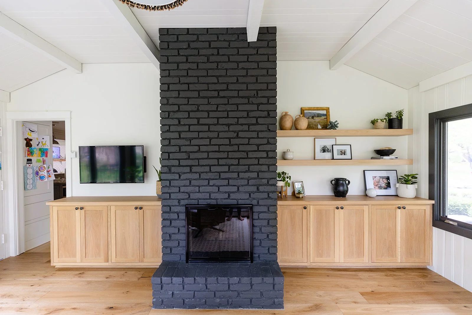 Living room with a black brick fireplace, wooden cabinets, and open shelves decorated with plants, framed photos, and vases; a wall-mounted TV is on the left side.