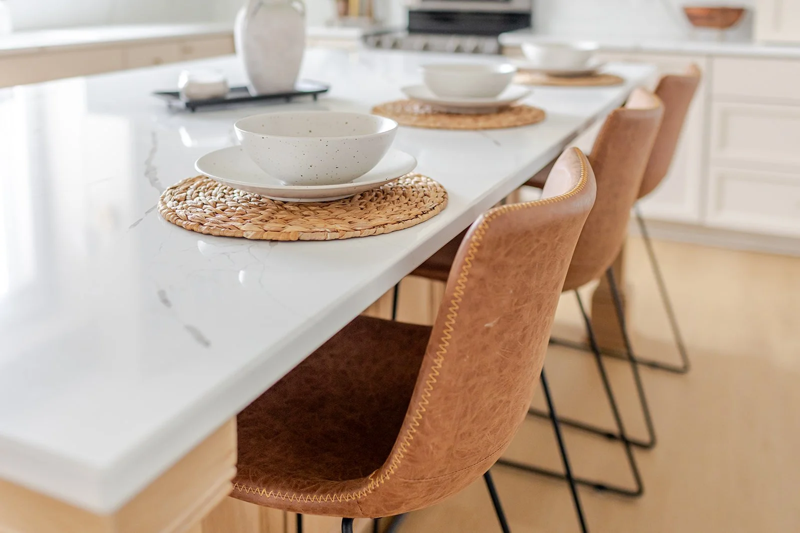 A white marble kitchen island with place settings of white bowls and plates on woven placemats, brown leather chairs with yellow trim, and a white background with cabinets.