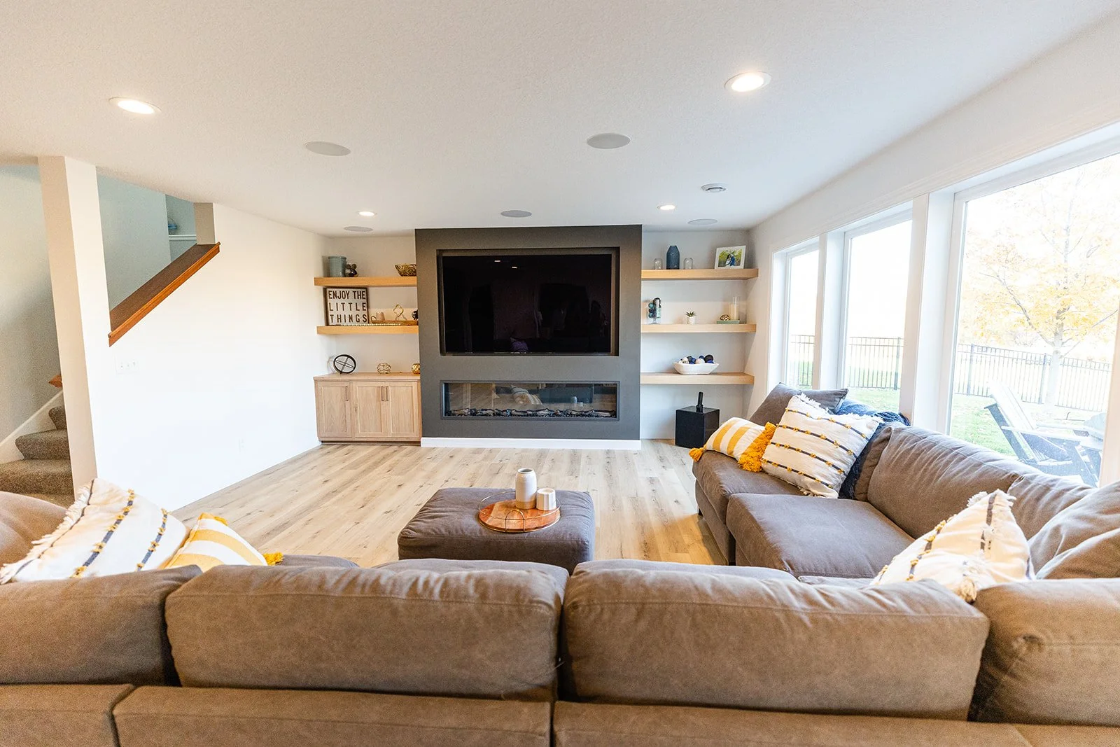 A living room with a large beige sectional sofa decorated with striped yellow and white pillows. A wooden coffee table with candles is in front of the sofa. The room features a flat-screen TV mounted above a modern fireplace on a gray accent wall, wi