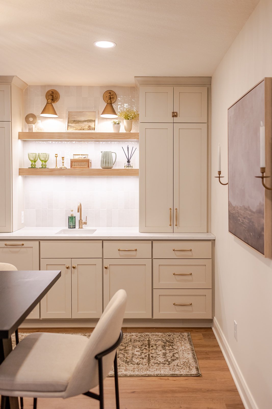 A modern kitchen with light beige cabinets, a white backsplash, and open wooden shelves displaying green glasses, candles, and decorative items. A gold faucet is above the sink, and a beige chair is partially visible in the foreground. A large painti