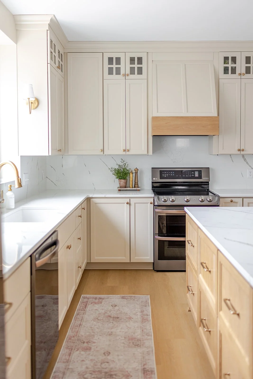 A modern kitchen with white cabinets, a stainless steel stove, a marble backsplash, a plant on the counter, and a pink patterned rug on wooden flooring.