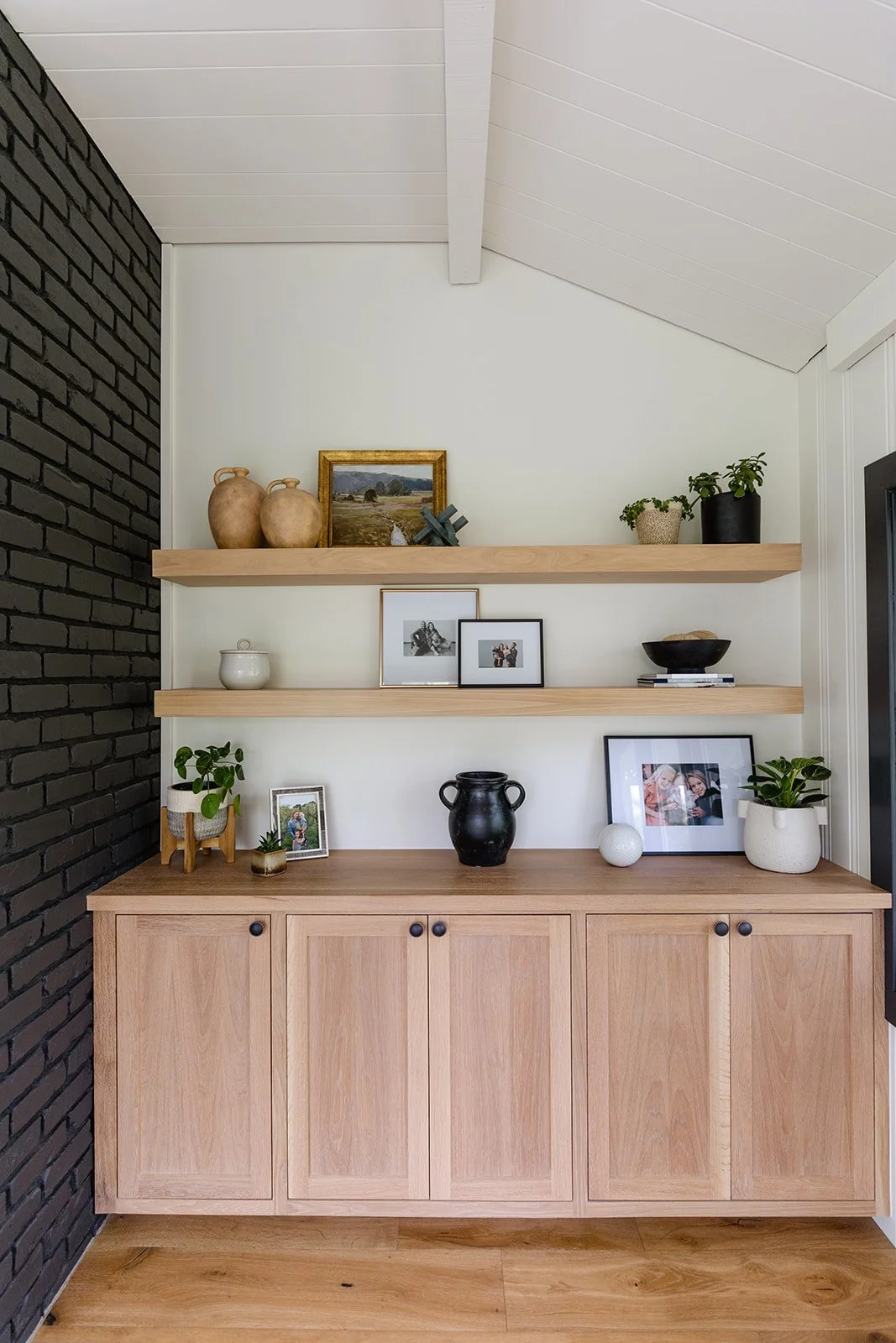 A wooden sideboard with black knobs under two floating shelves decorated with potted plants, framed photographs, pottery, a bowl, and decorative objects in a modern living space with a black brick wall on the left and white ceiling and walls.