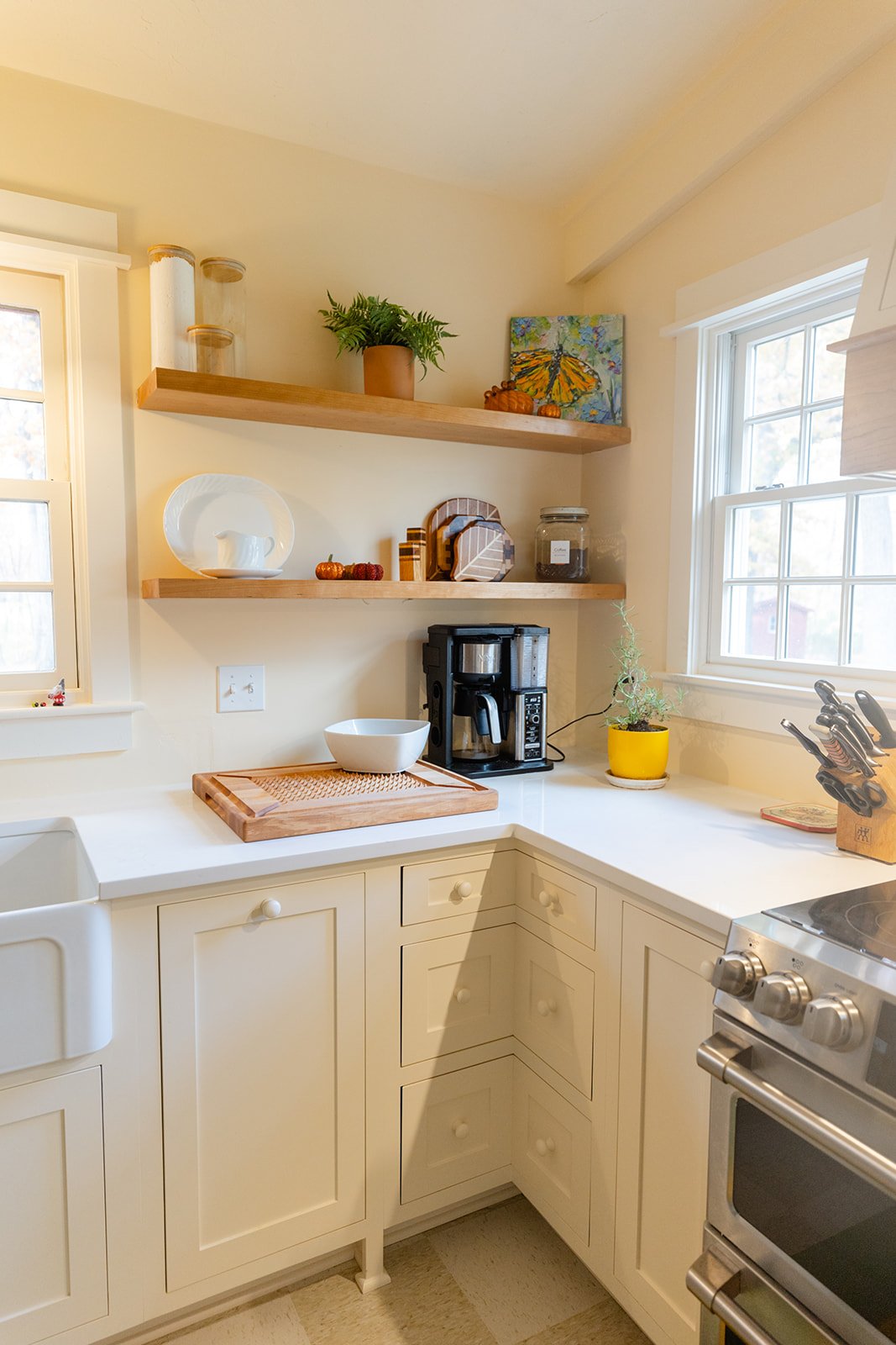 Kitchen with white cabinets, two wooden shelves decorated with pumpkins, a potted fern, art, and kitchen appliances including a coffee maker and a stainless steel stove.