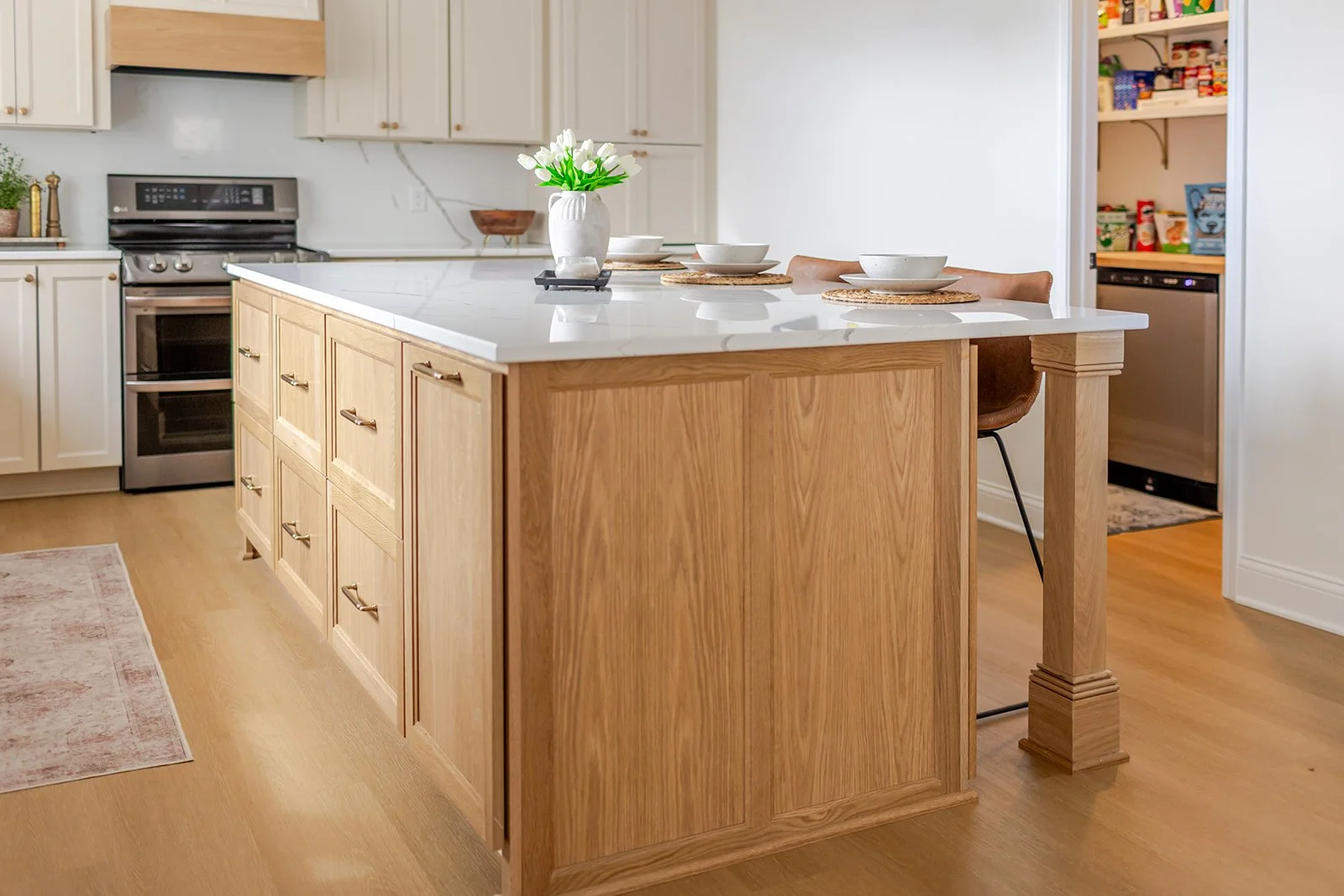 Kitchen with an island, white cabinets, wood drawers, and a white countertop. Decor includes a vase with white flowers, bowls, and placemats. In the background, a stove, a small potted plant, and an open pantry with snacks are visible.