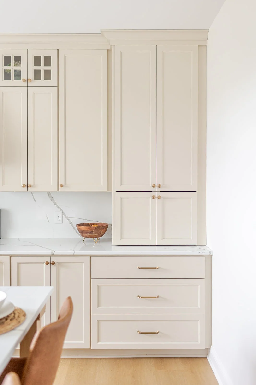 Kitchen cabinet with cream-colored drawers and cabinets, marble countertop, and a wooden bowl with fruit.