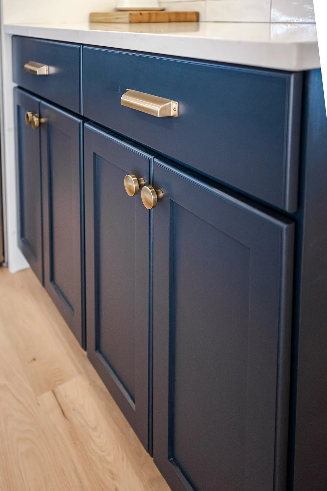 A navy blue kitchen cabinet with brass handles and knobs, topped with a white countertop, with a wooden cutting board on top.
