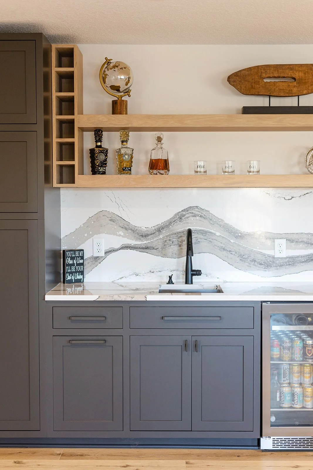 Kitchen with gray cabinets, a black faucet, marble countertop, and open wooden shelves holding decorative bottles, glasses, and a globe sculpture.