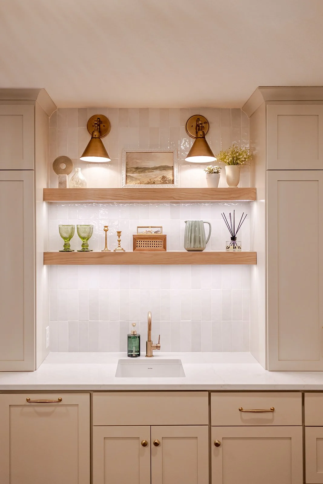 Kitchen sink with beige cabinets, a white countertop, and two wooden open shelves above holding decorative items. Two brass wall-mounted lights illuminate the shelf area, which includes plants, dishes, candles, and a framed painting.
