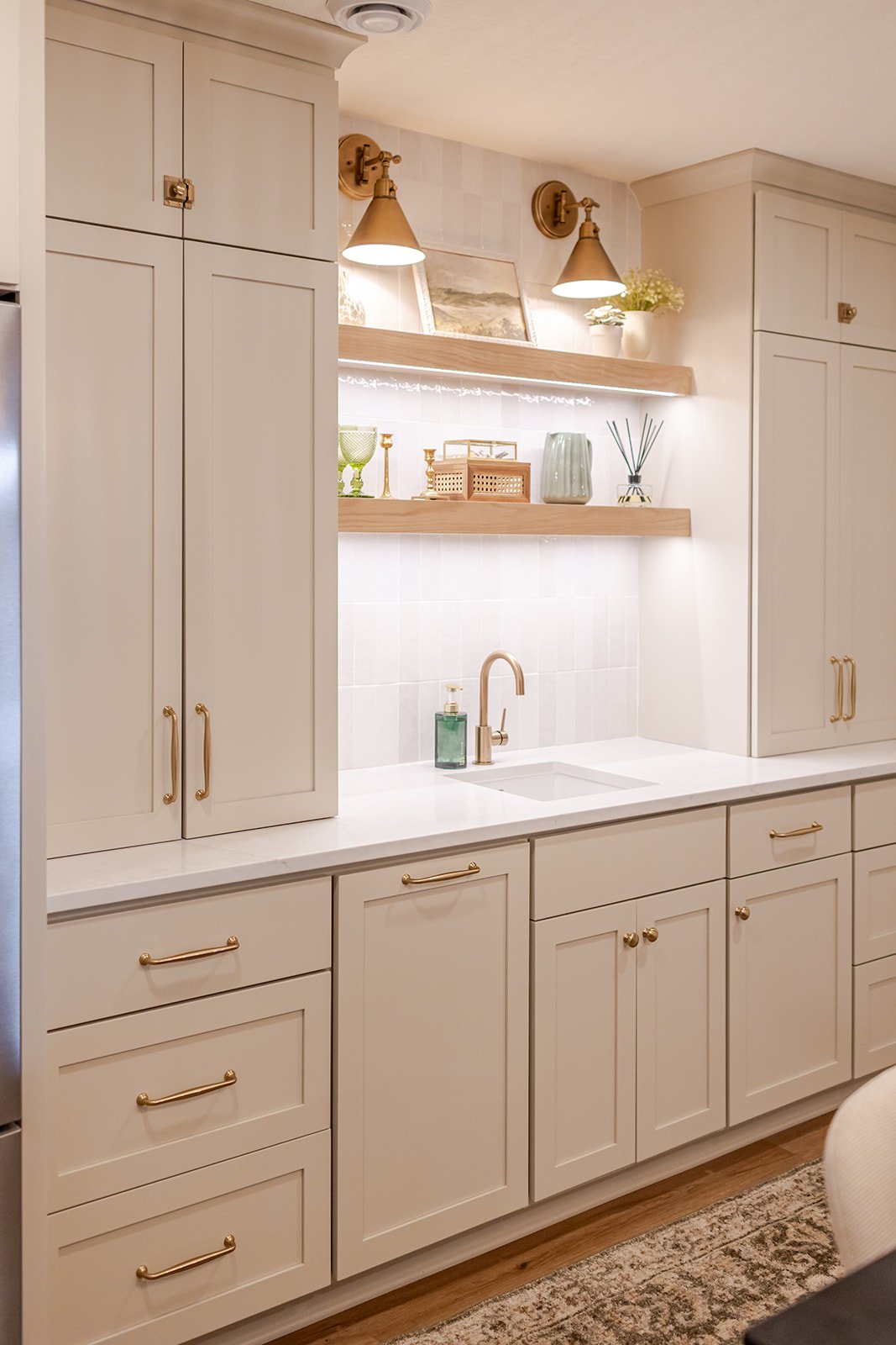 Cream-colored kitchen with gold handles, gold wall sconces, wooden shelves, and decorative items including a potted plant, vases, and framed artwork.