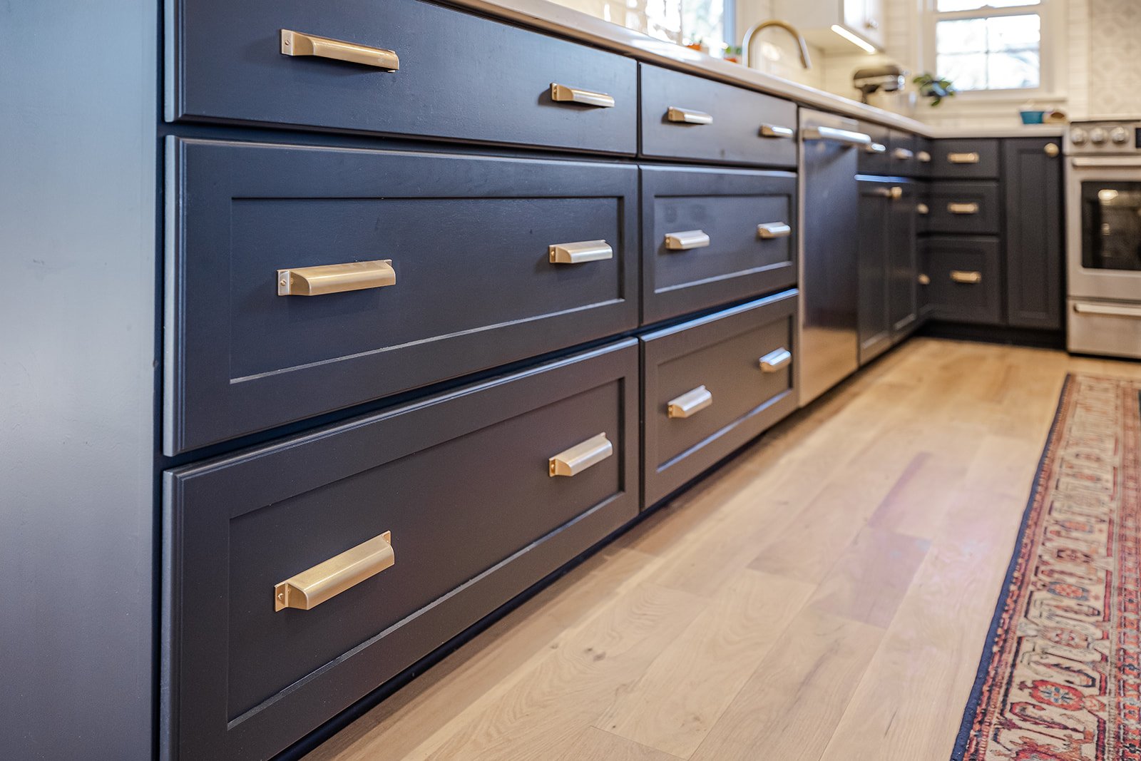 Close-up view of dark blue kitchen cabinets with gold handles, wooden floor, and part of a patterned rug visible at the bottom right, with kitchen appliances in the background.