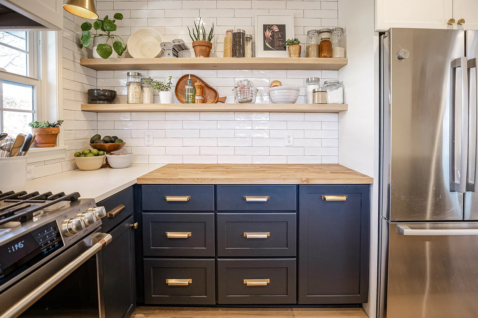 Kitchen with navy cabinets, wooden countertops, open shelves with jars, bowls, plants, framed art, and a window with sunlight, next to a stainless steel refrigerator and a stove.