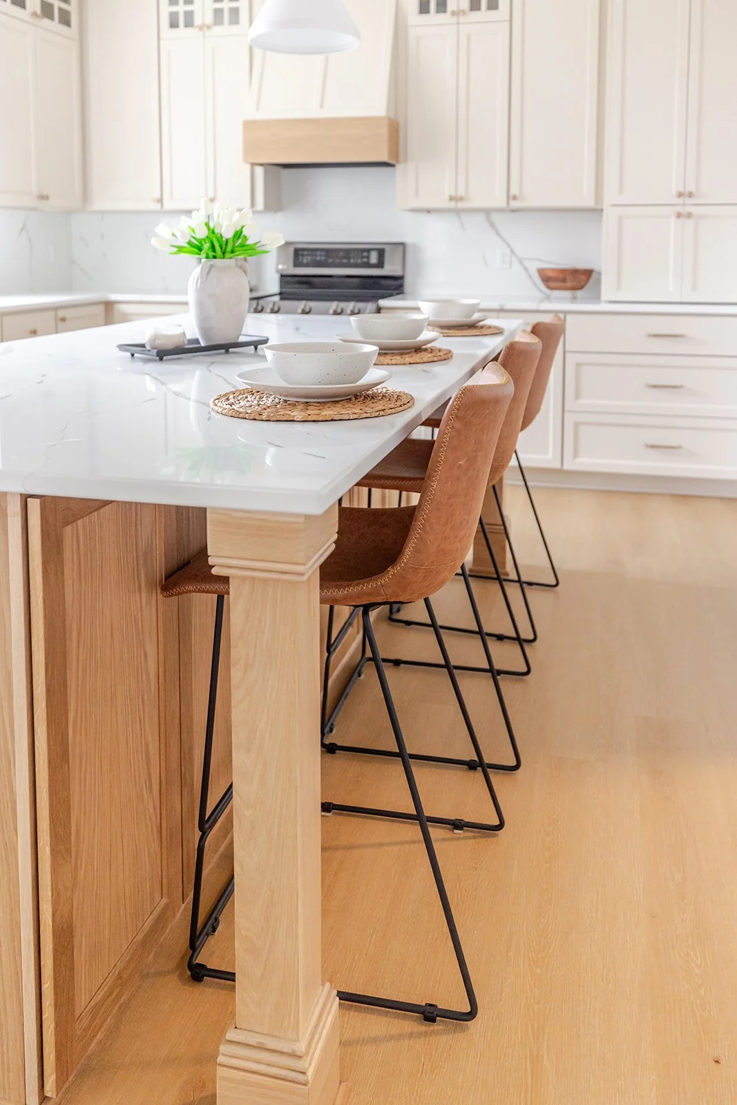 Modern kitchen with white cabinetry, white marble countertops, and wooden accents. A white island has four brown leather bar stools with black metal legs. On the island are white bowls on woven placemats and a white vase with white tulips.