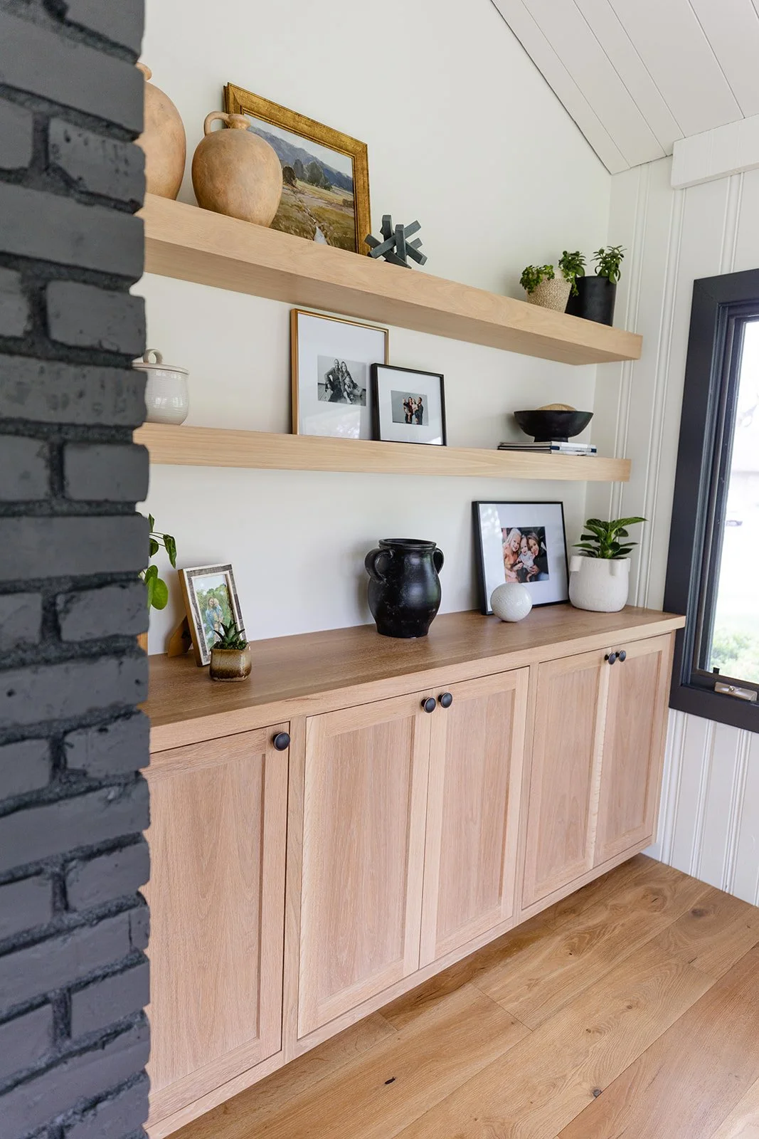 A wooden sideboard with black knobs decorated with potted plants, framed photos, and vases; two open shelves above with vases, artwork, and decor; window to the right, partly open; adjacent black brick wall on left side.