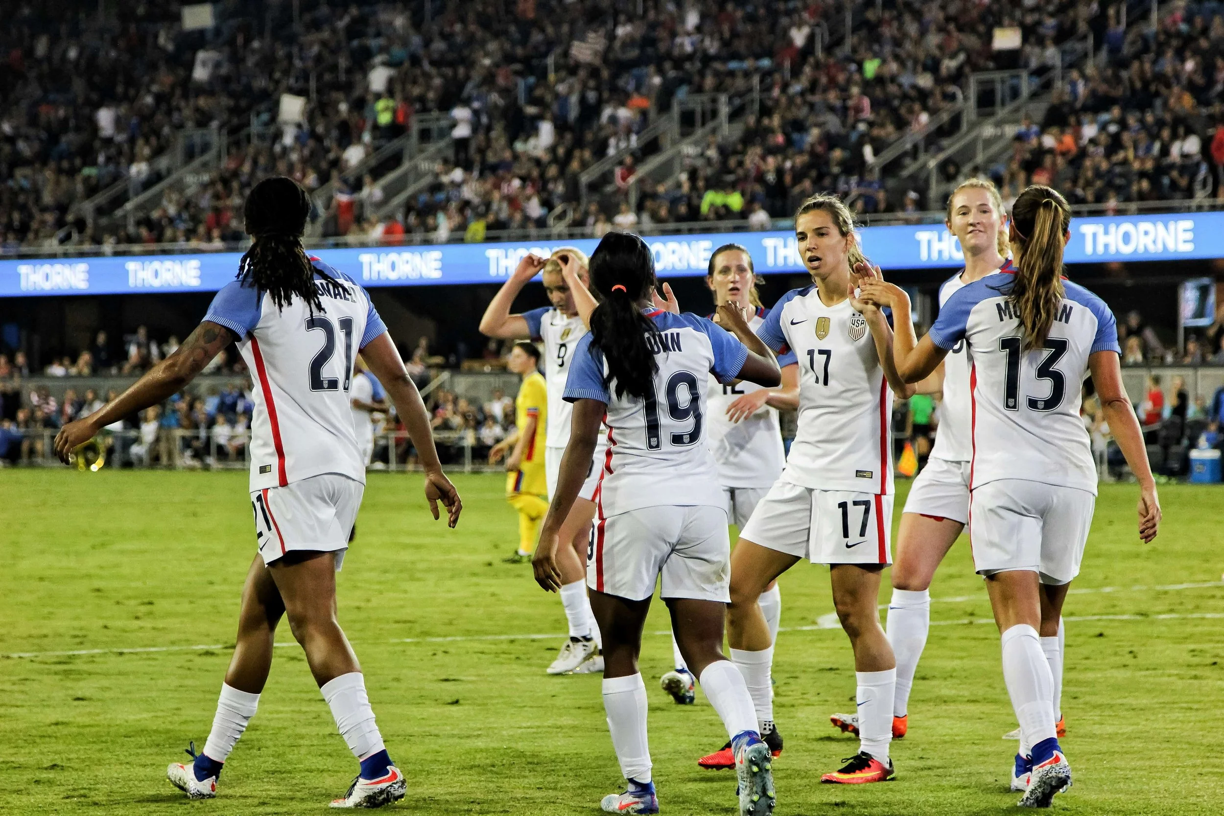 Women’s soccer team on the field, wearing white jerseys with blue sleeves and red and blue accents, celebrating during a match in a stadium filled with spectators.