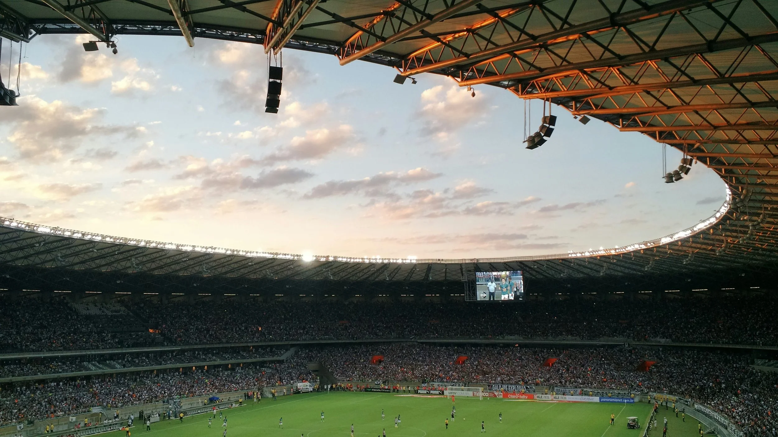 A large soccer stadium filled with spectators, with a game in progress on the field. The stadium has a partially open roof with visible support beams and hanging speakers, and the sky shows a sunset with scattered clouds.