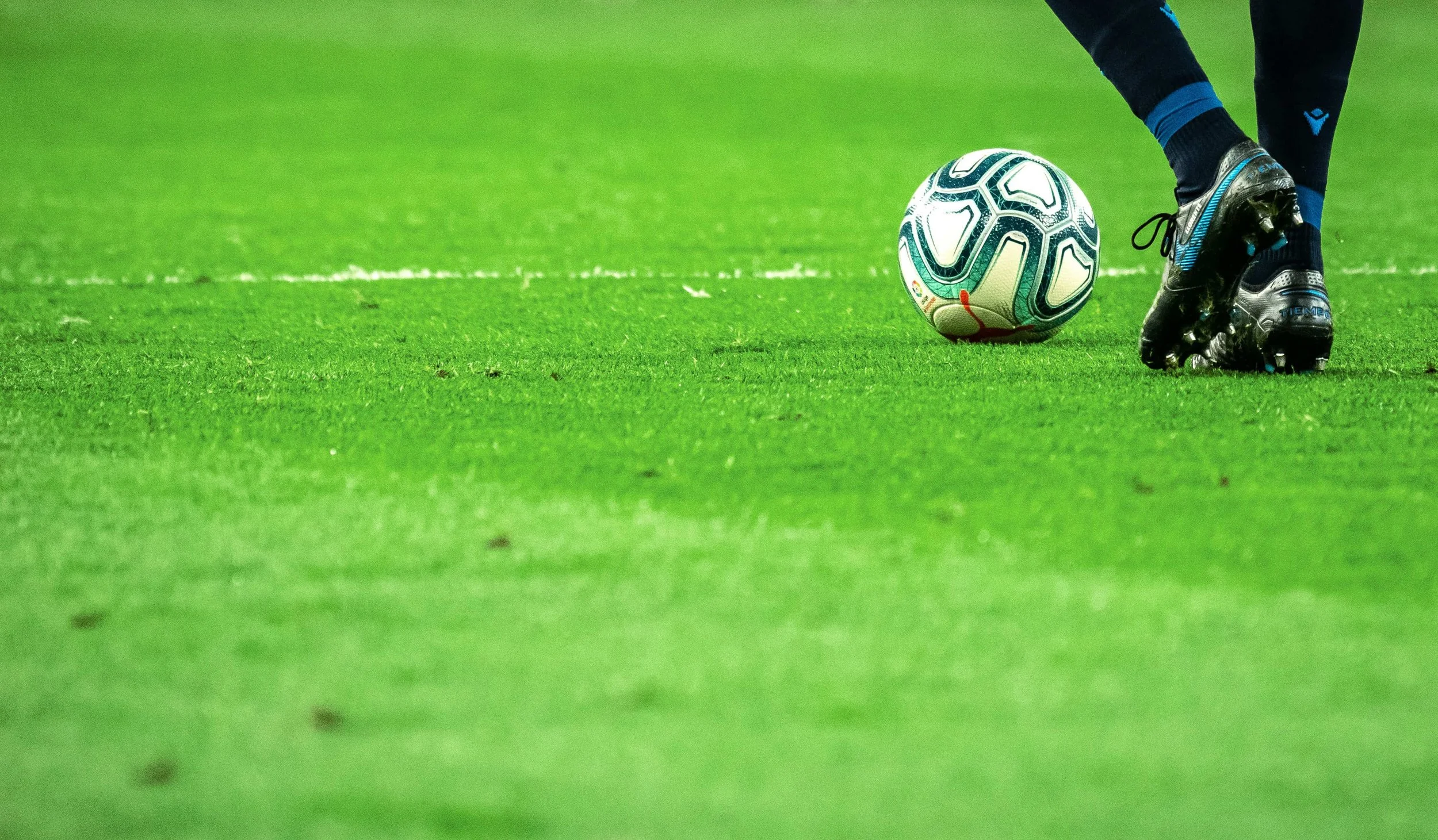 Close-up of a soccer player's feet in black shoes and blue socks preparing to kick a soccer ball on a green field.