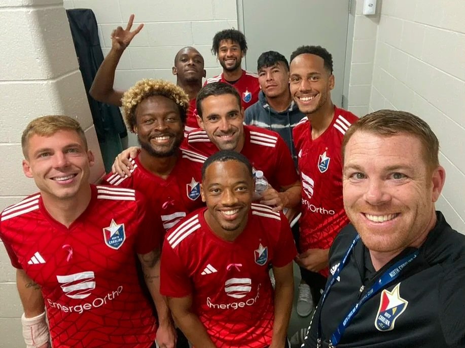 Group of soccer players and a coach taking a selfie in a locker room, with some players making peace signs and smiling.