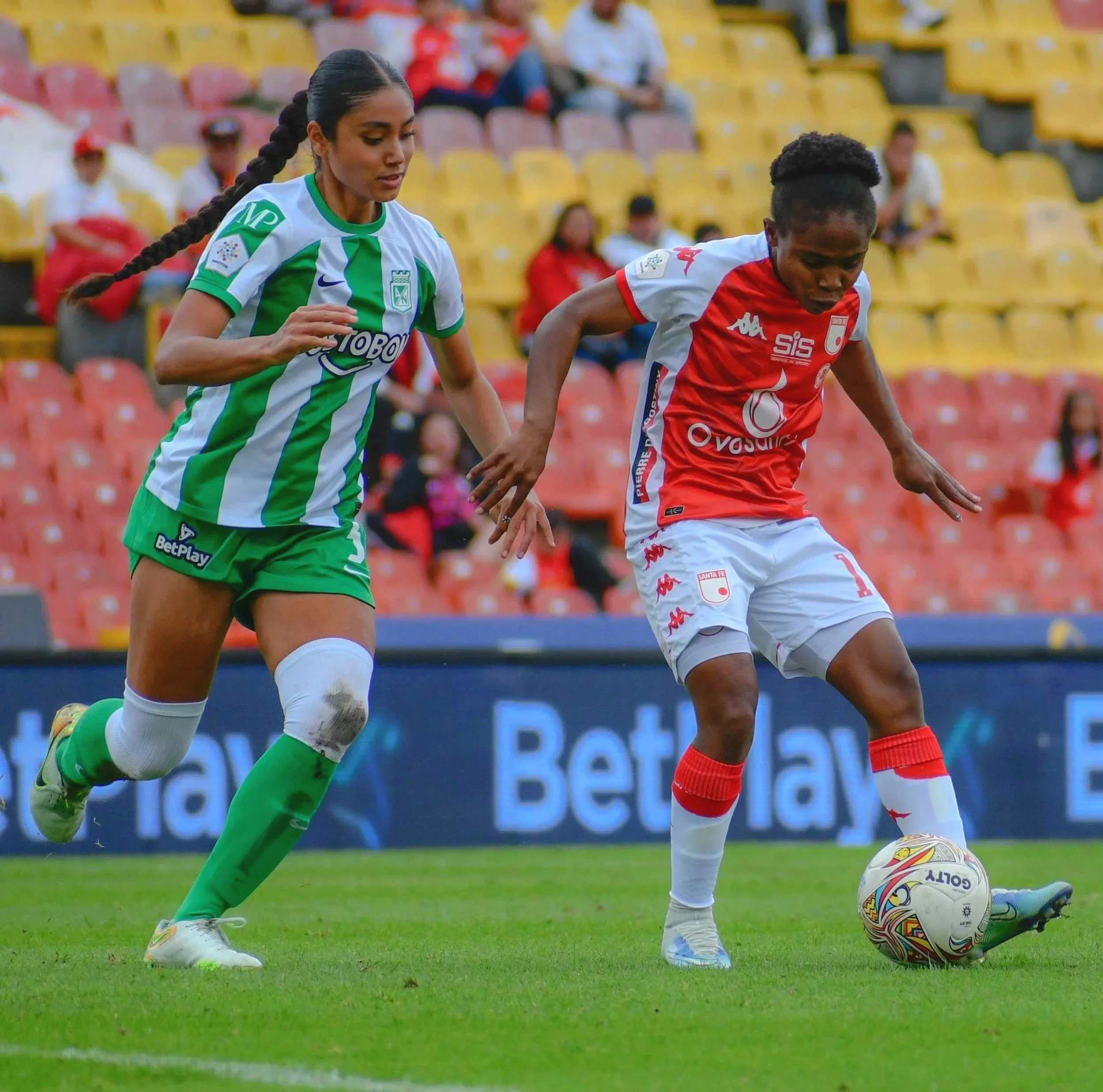 Two women playing soccer on the field, with one in a green and white uniform and the other in a red and white uniform, with the player in red controlling the ball.