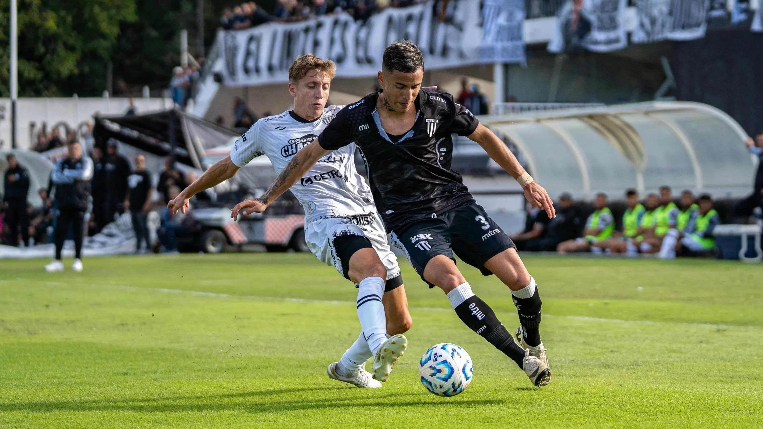 Two soccer players compete for the ball on a grassy field during a match. One player in black jersey and black shorts is in front, while the other in white jersey and shorts is behind him. Several spectators and team staff are visible in the background.