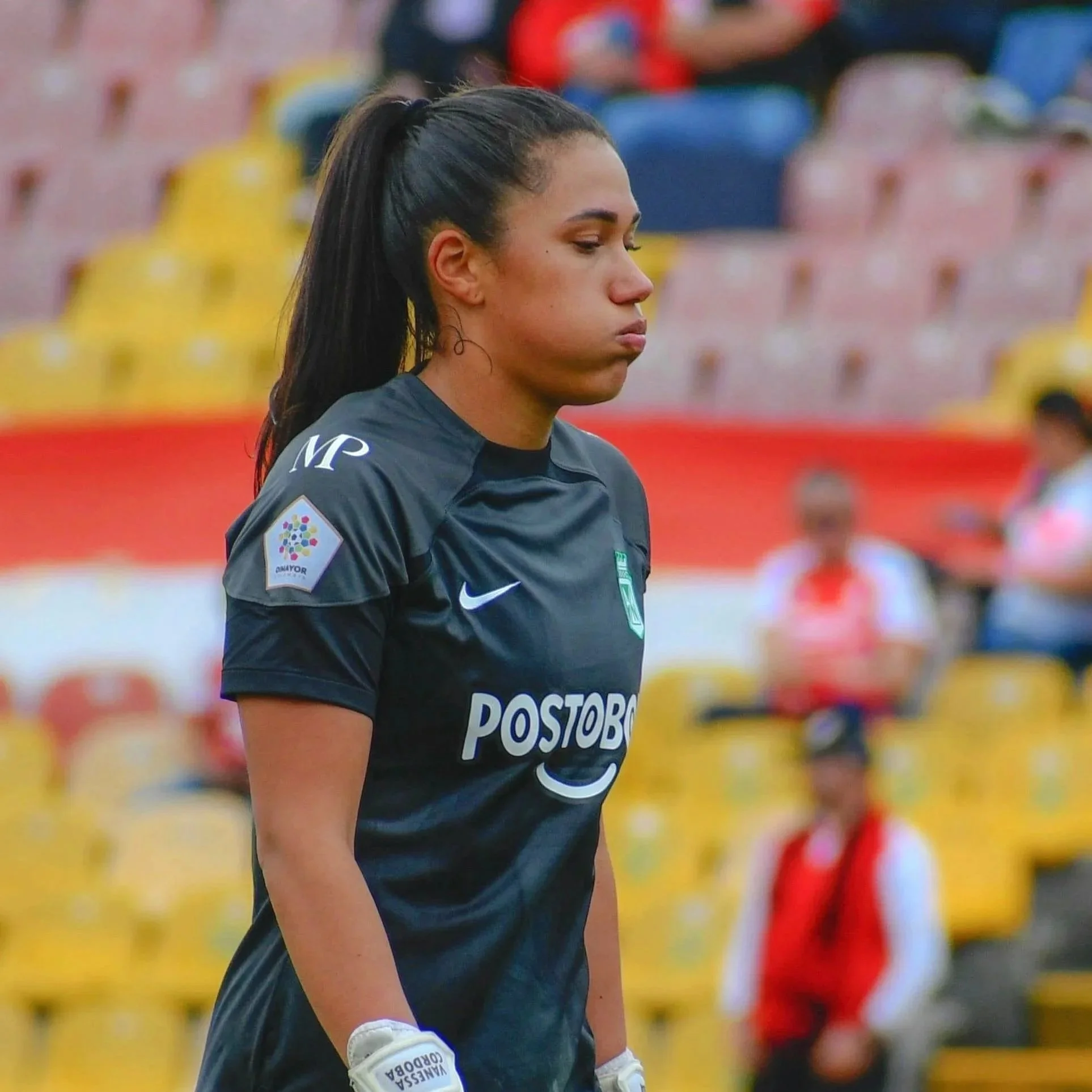 A female soccer player standing on the field wearing a dark blue uniform with white branding, including gloves, with a serious expression.