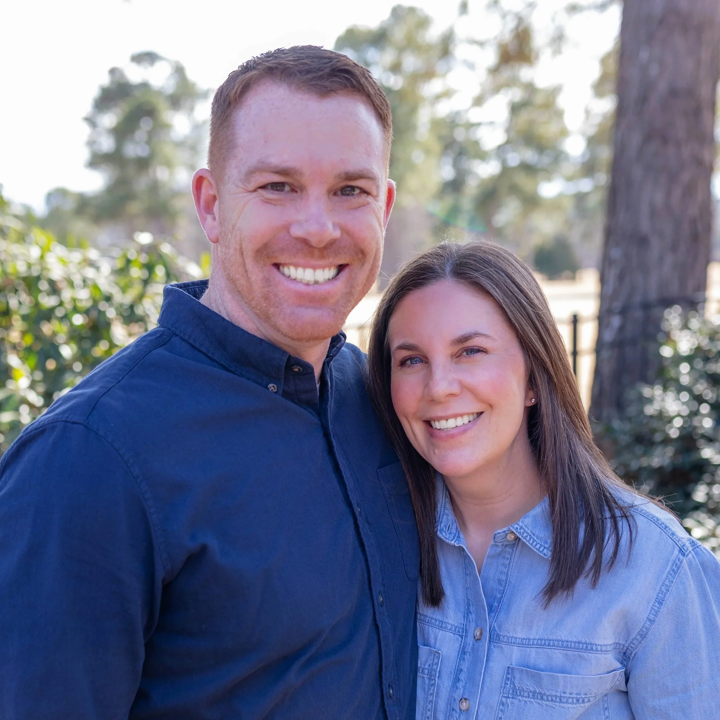 A smiling man and woman standing outside, with trees and sunlight in the background.
