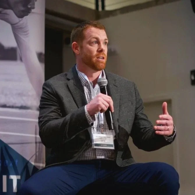 A man in a gray blazer, checkered shirt, and dark pants sitting on a chair speaking into a microphone at an event.