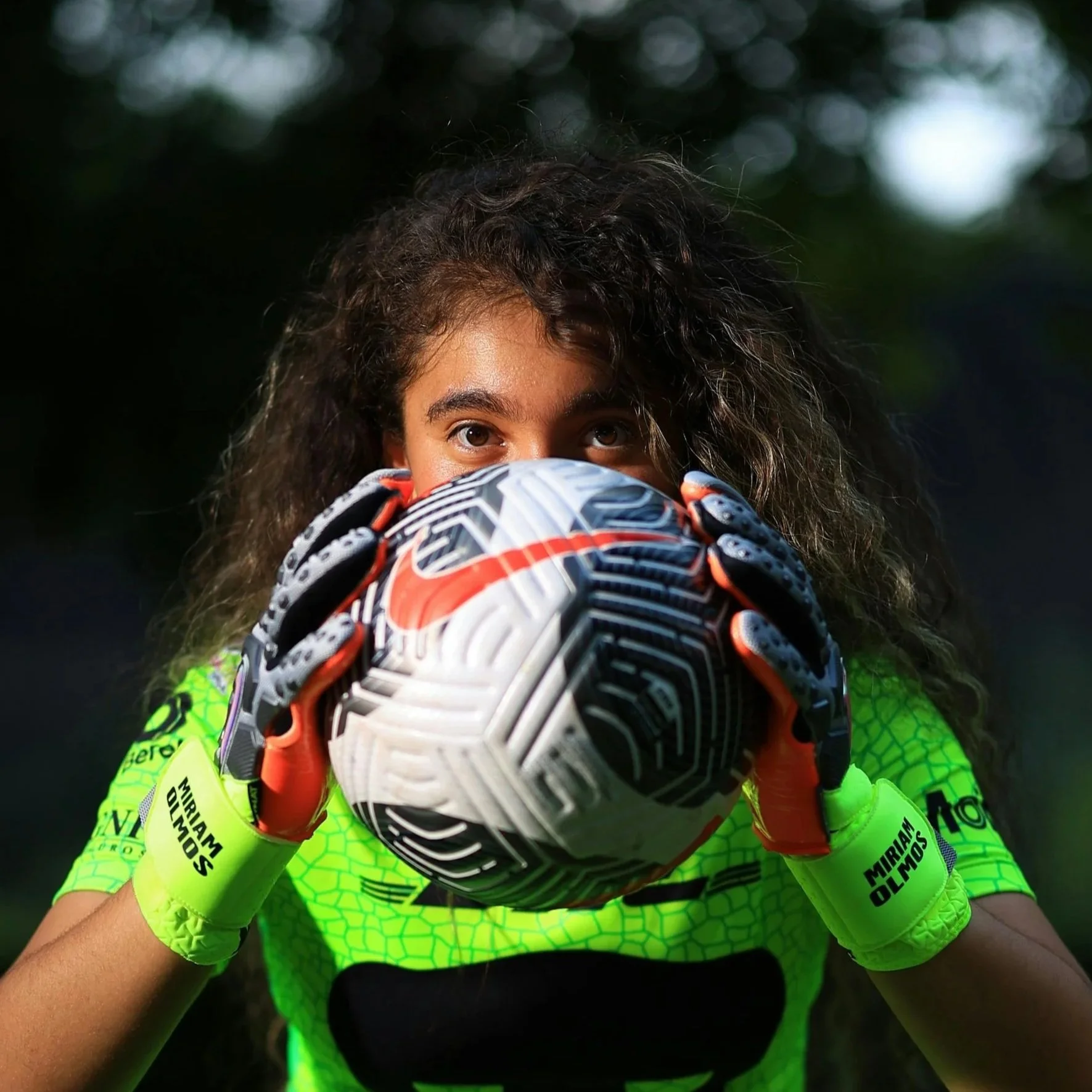 Young girl wearing a neon yellow sports jersey and gloves, holding a patterned soccer ball close to her face, with a blurred dark outdoor background.