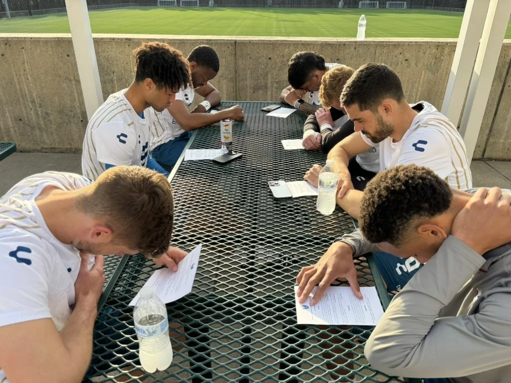 Several young men sitting around a metal outdoor table, reading papers and drinking water, on a sports field.