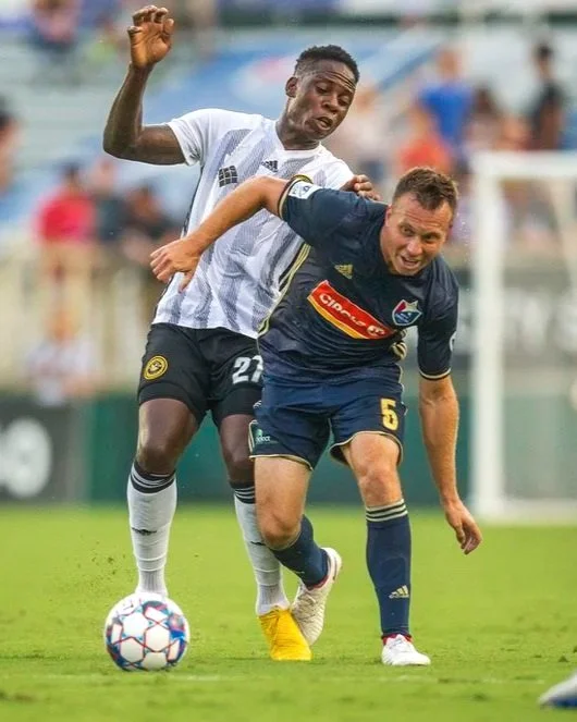 Two soccer players competing for the ball during a match, one in a white and black uniform and the other in a dark blue uniform, on a grassy field with a blurred crowd in the background.