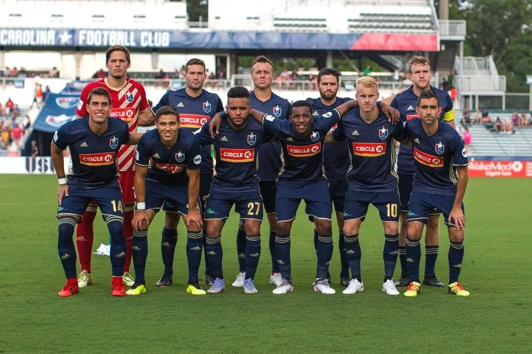 Soccer team in blue jerseys and shorts posing on a field, with a stadium and fans in the background.