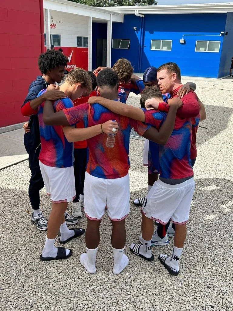 A group of young athletes, likely a soccer team, huddles together with their arms around each other outdoors near a colorful building.
