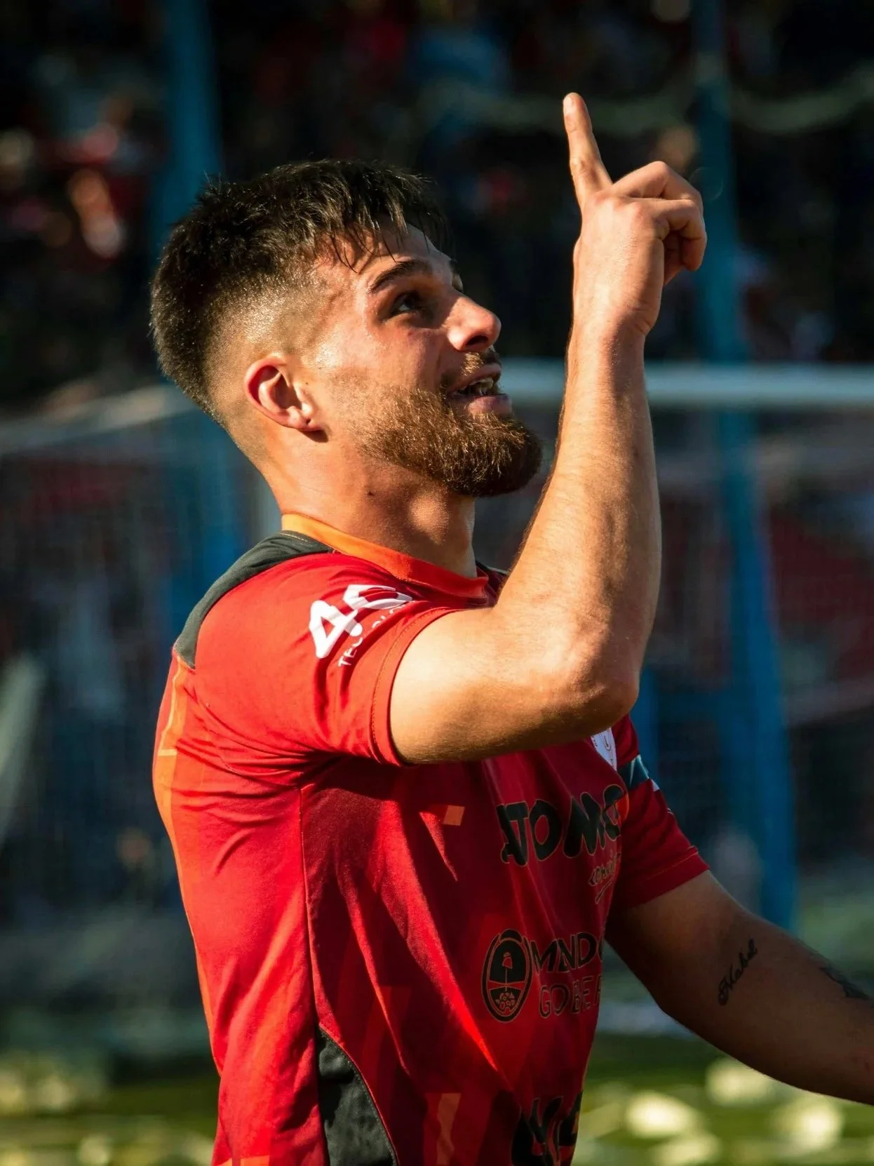 A soccer player with a beard and short hair, wearing a red jersey, pointing upward, outdoors on a sunny day.