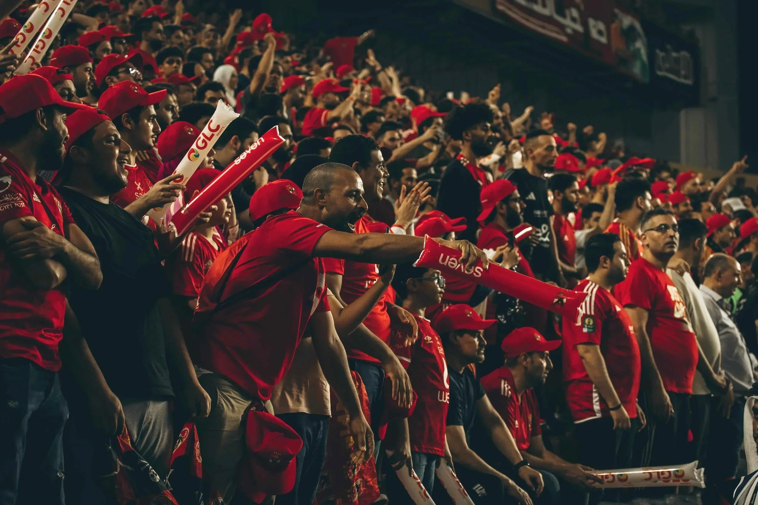 Crowd of sports fans dressed in red, cheering and holding inflatable cheering sticks at a stadium.