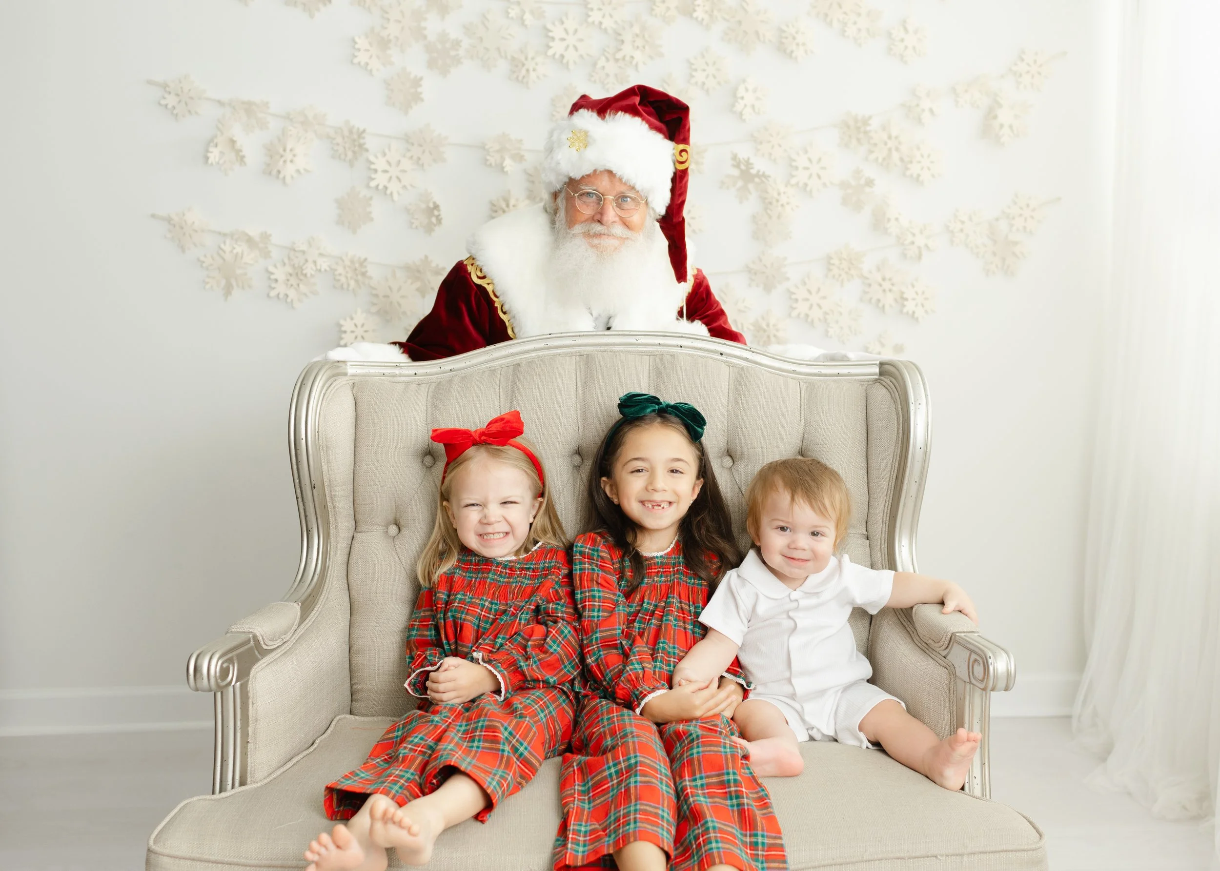 Three siblings in coordinated Christmas outfits smiling as Santa peeks over the settee
