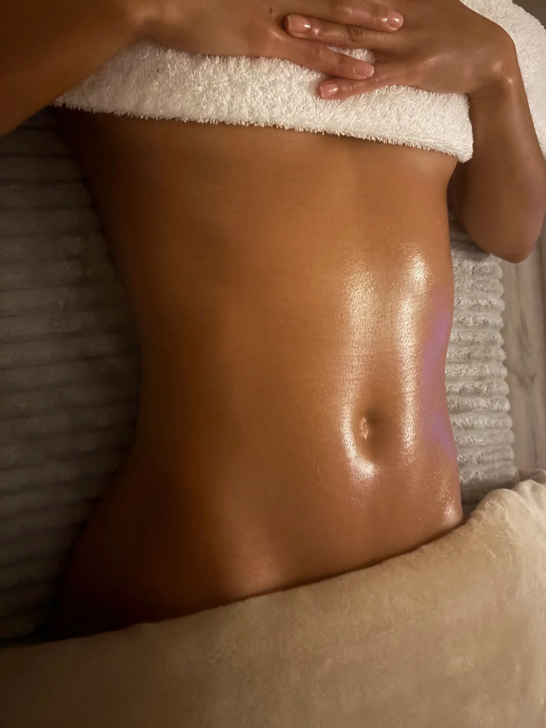 Close-up of a woman's toned, shiny abdomen with a belly button, partially covered by a white towel, lying on a striped textured surface.