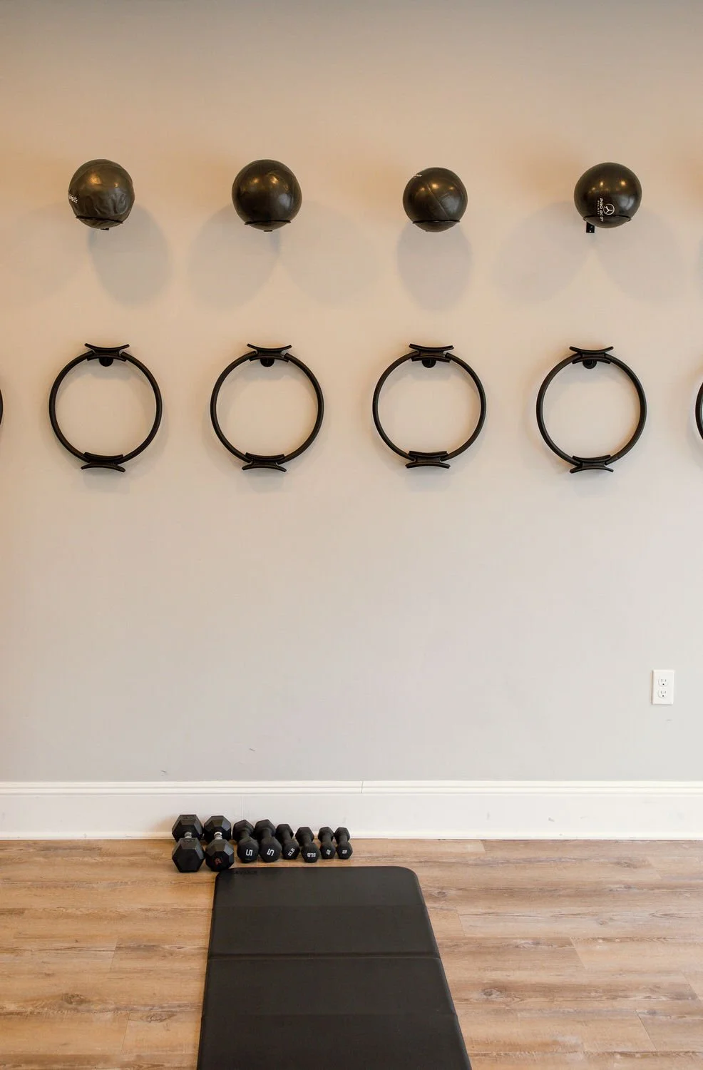 Wall with four black medicine balls mounted above four black workout rings, weight dumbbells on the floor, and a black exercise mat in front at Lava Fitness, Lavallette NJ.