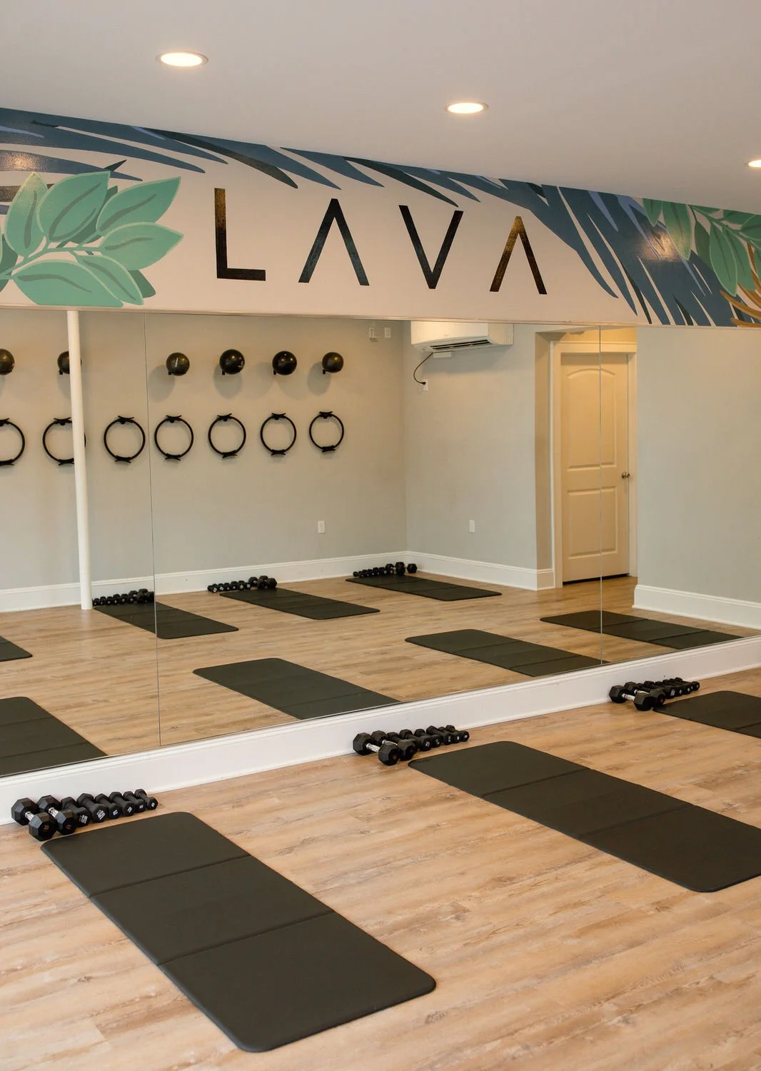 Fitness room with mirrors, black yoga mats, dumbbells, wall-mounted kettlebells, and decorative wall art with greenery and the word 'LAVA' at Lava Fitness, Lavallette NJ.